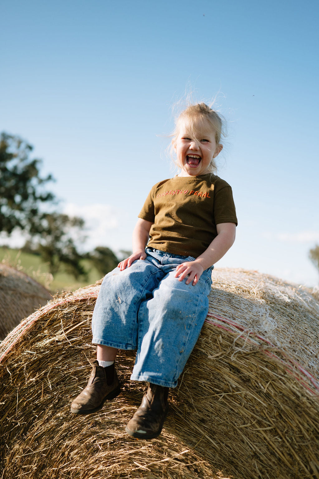 Laughing child in the brown CURRENTLY FERAL kids t-shirt sitting on a hay bale