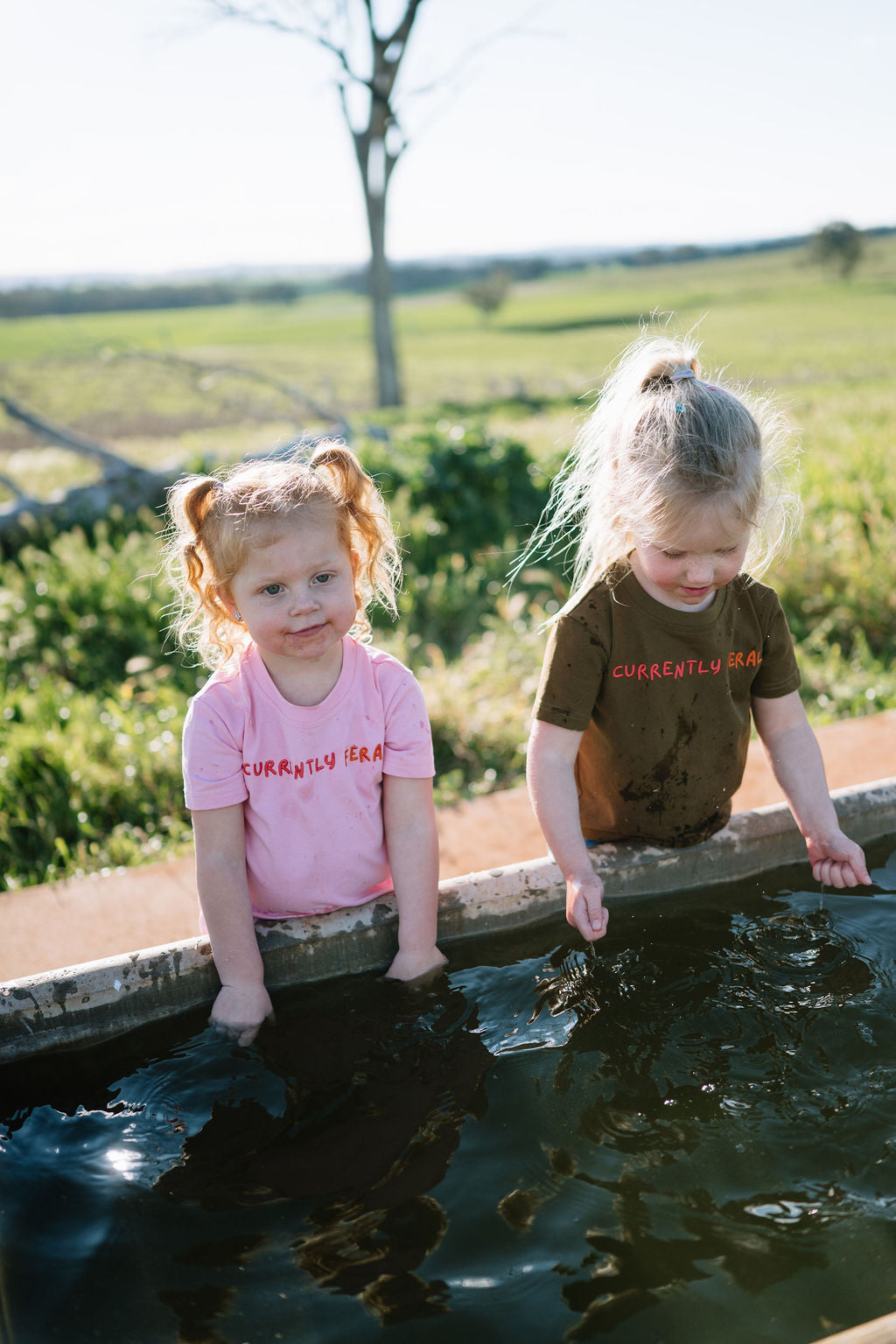 Two children at a farm water trough in pink and brown CURRENTLY FERAL kids t-shirts