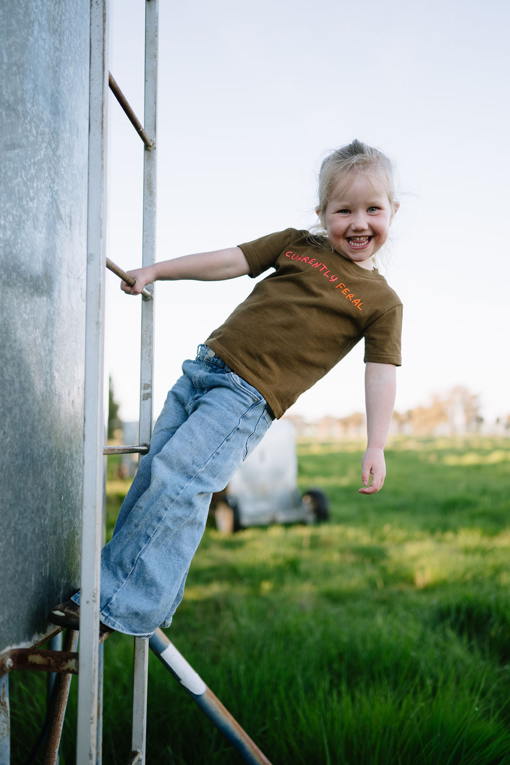 Child in the brown CURRENTLY FERAL kids t-shirt climbing a ladder on the farm
