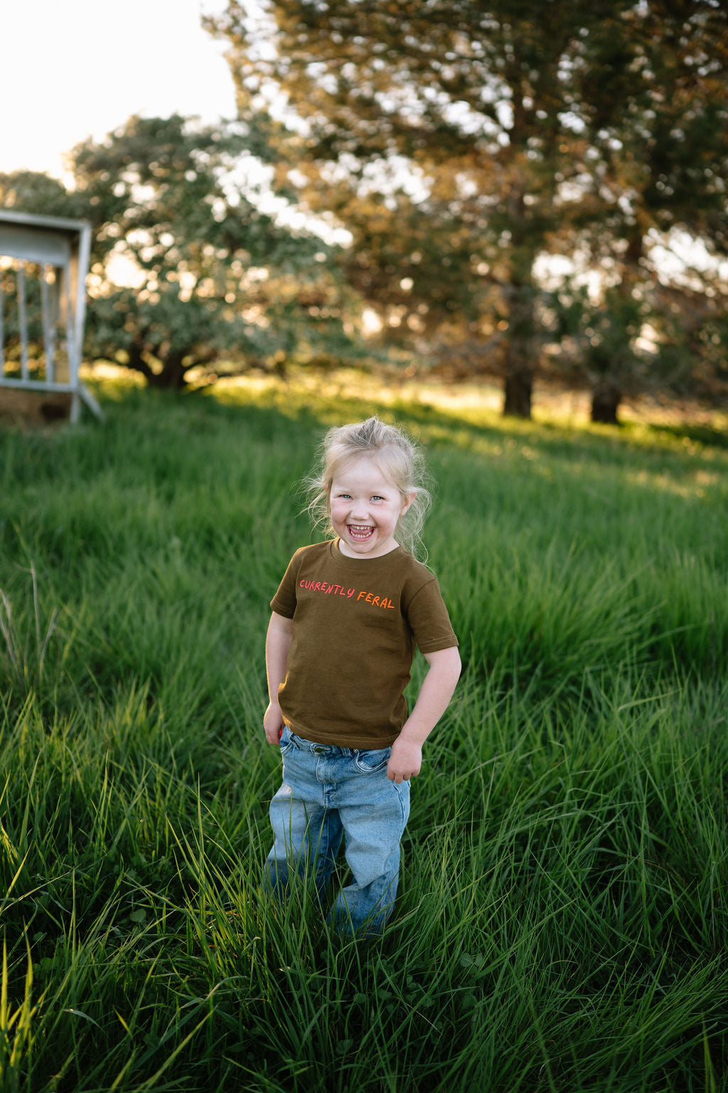 Child in the brown CURRENTLY FERAL kids t-shirt standing in green grass