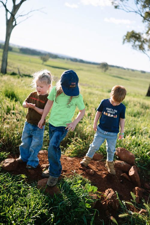 Three kids on red dirt in a green paddock wearing a green TEAM COLLIE tee, a brown CURRENTLY FERAL tee and a navy WHAT'S FOR SMOKO? tee