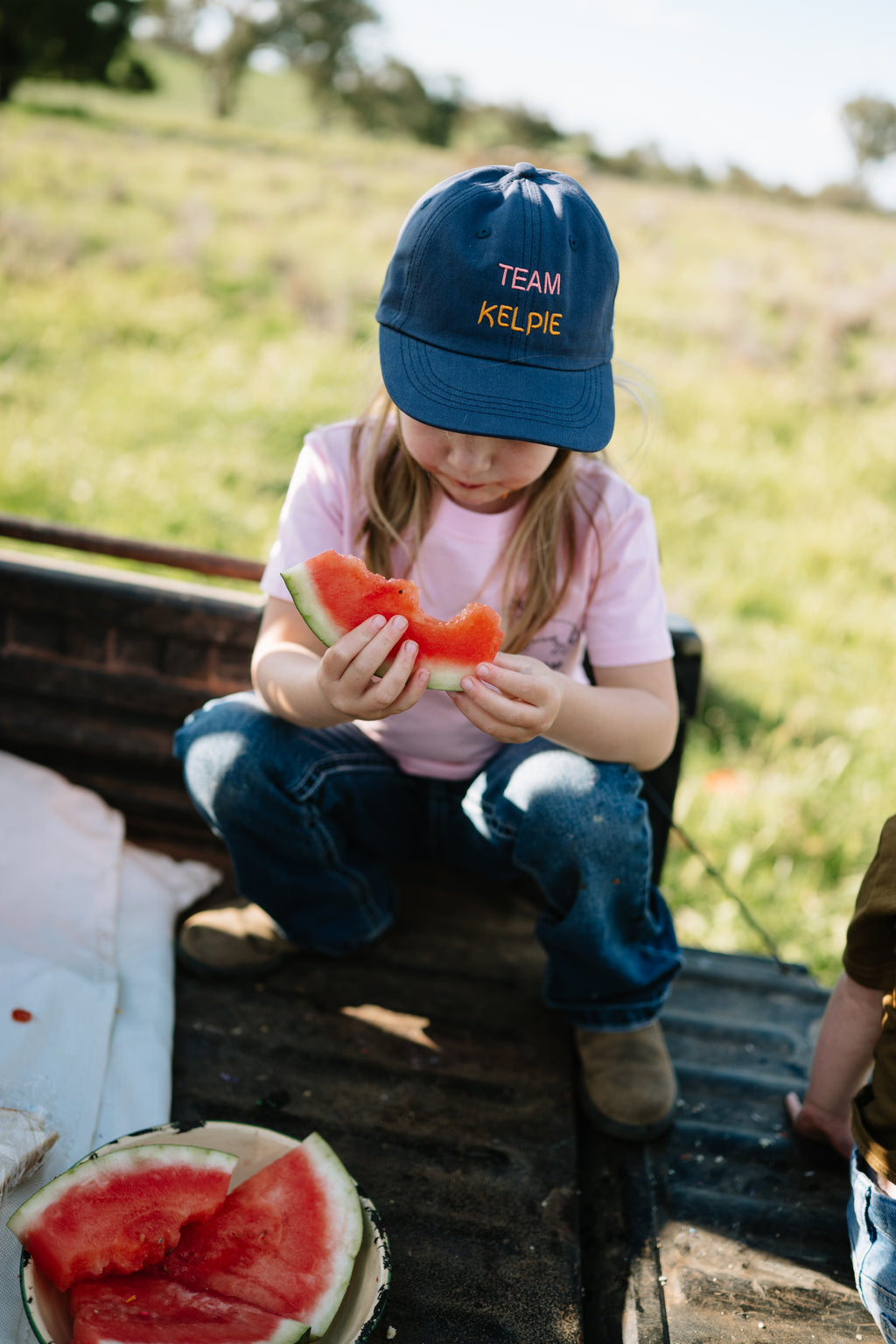 Child in the navy TEAM KELPIE kids cap eating watermelon on the back of a ute
