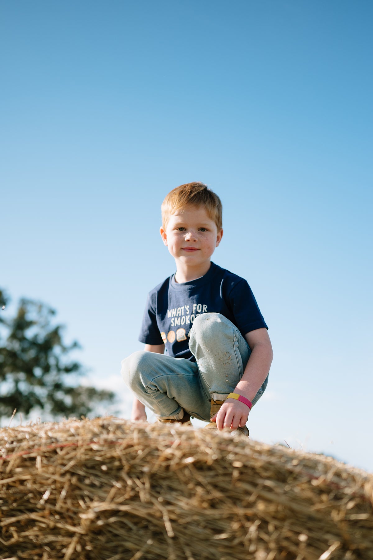 Child in the navy WHAT'S FOR SMOKO? kids t-shirt crouching on top of a hay bale
