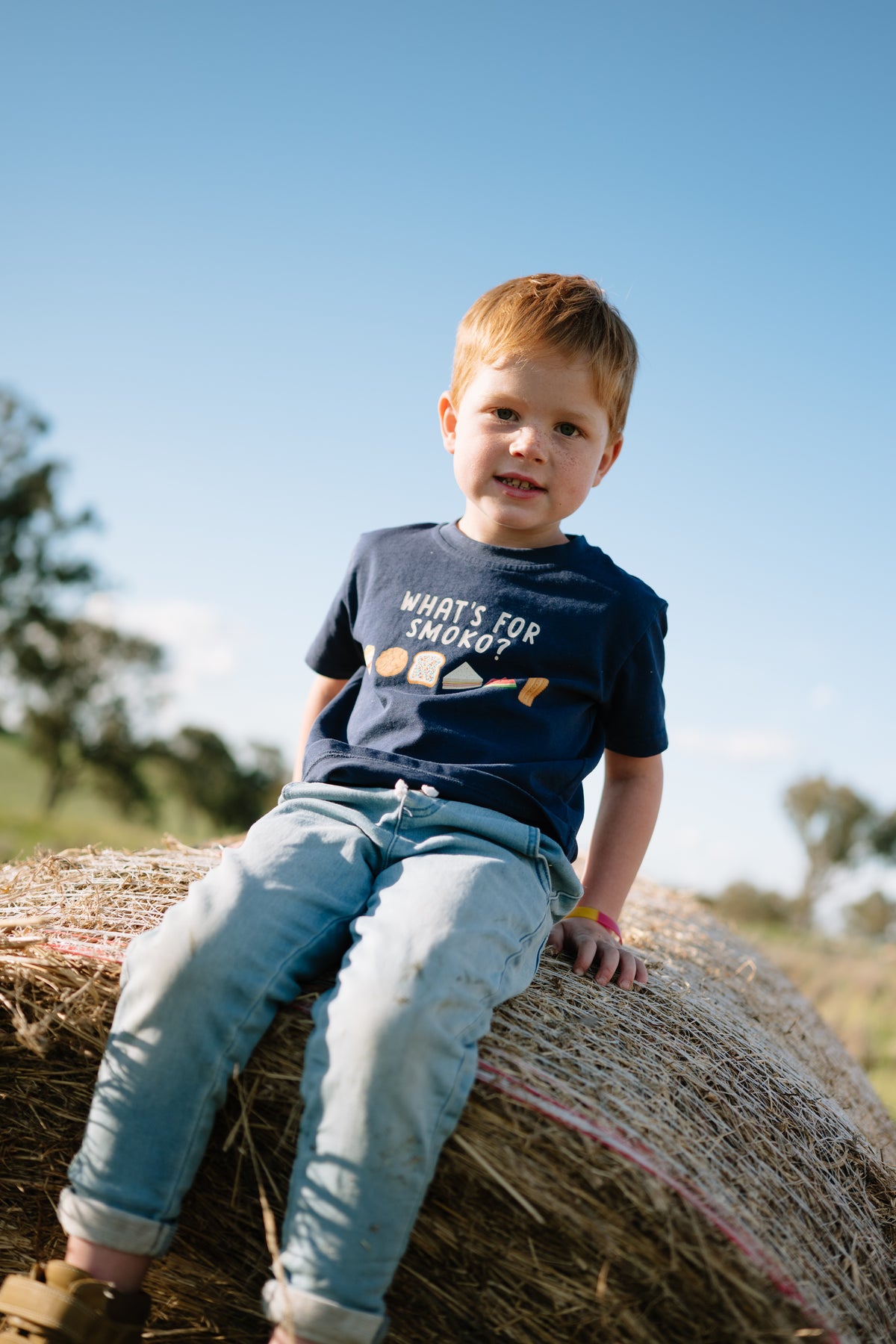 Child in the WHAT'S FOR SMOKO? kids t-shirt and jeans sitting on a round hay bale