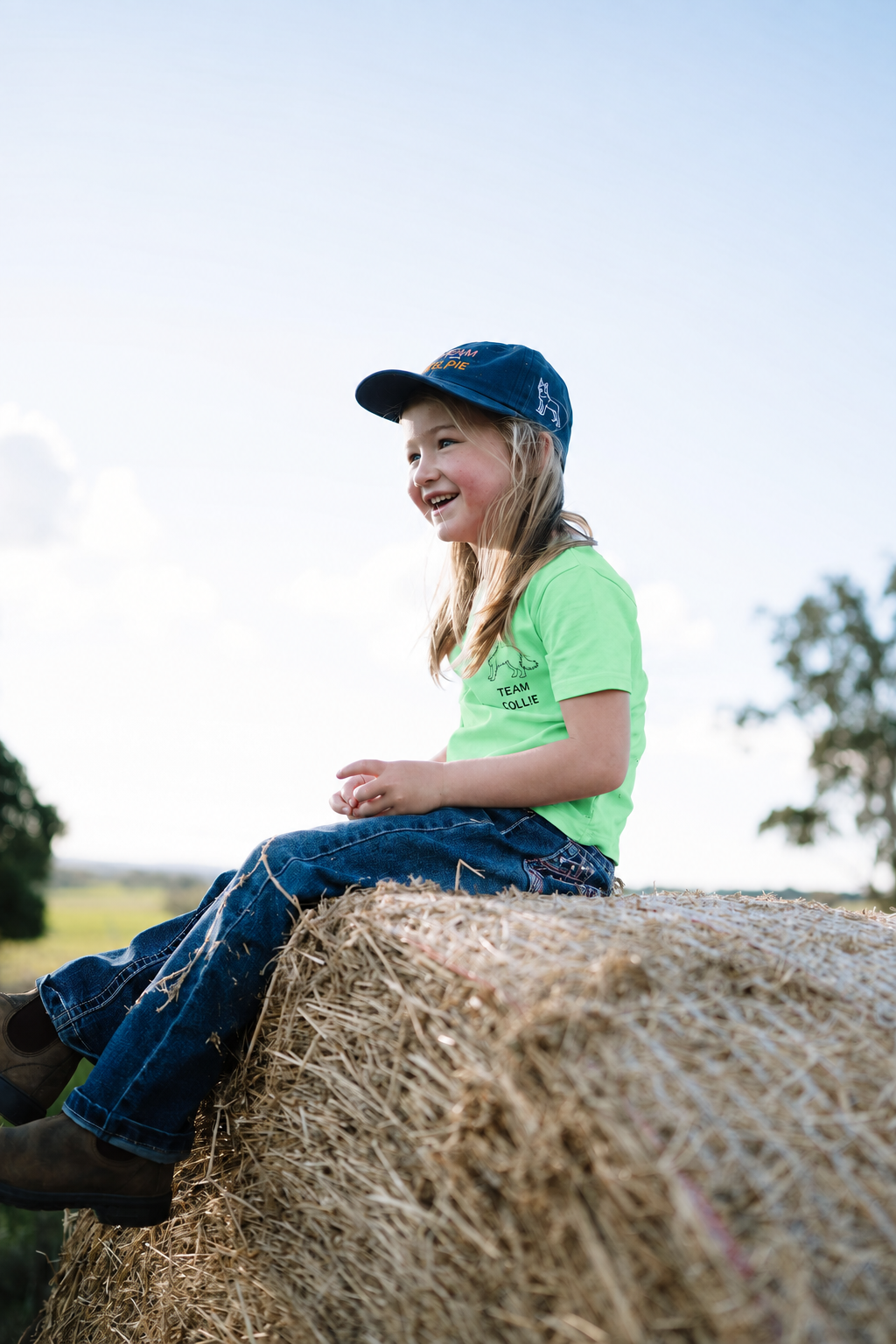 Smiling girl sitting on a hay bale in a green TEAM COLLIE kids t-shirt with a Border Collie embroidered on the chest